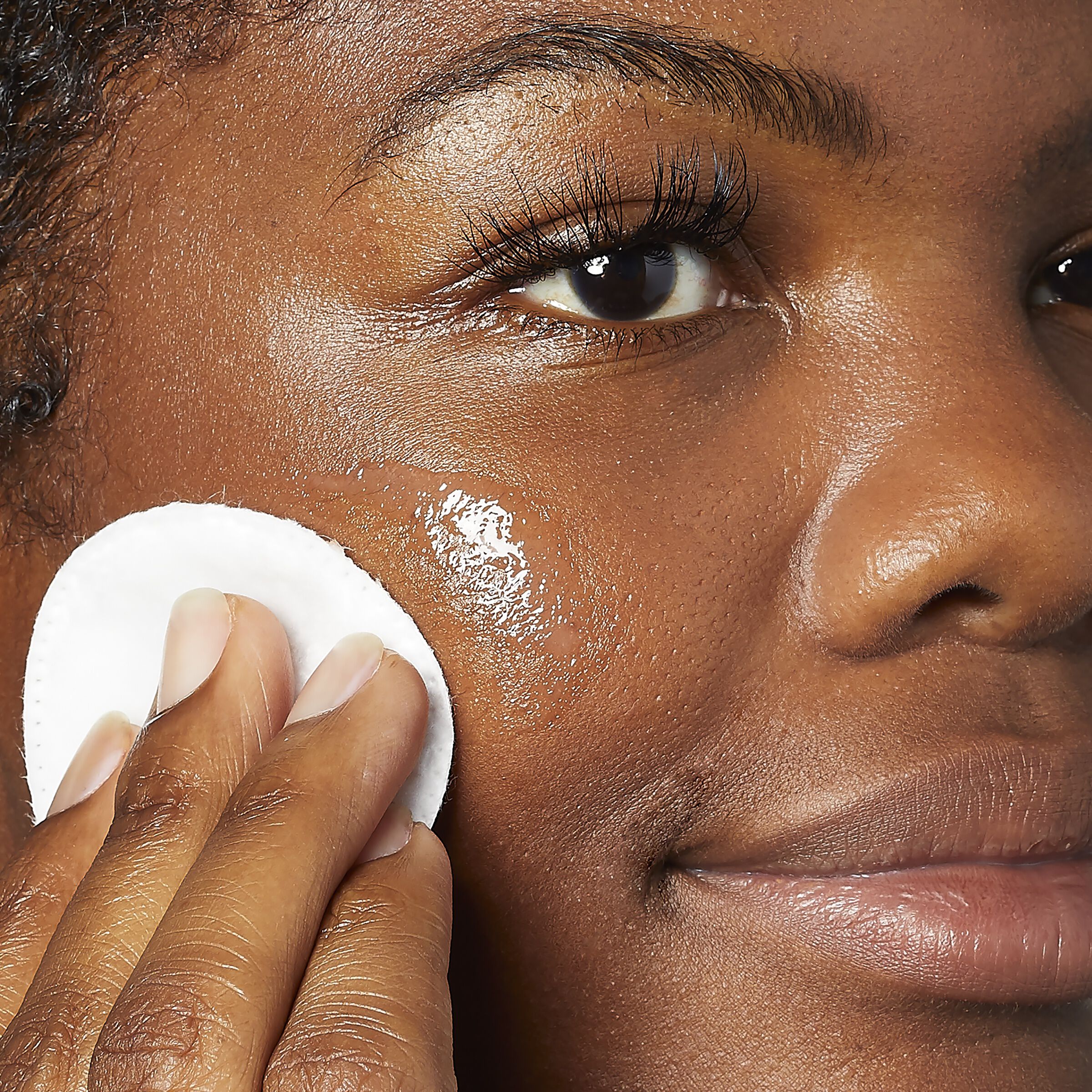 Close-up of a person gently applying skincare with a cotton pad to a shiny, moisturized cheek. The scene conveys a sense of calm and self-care.