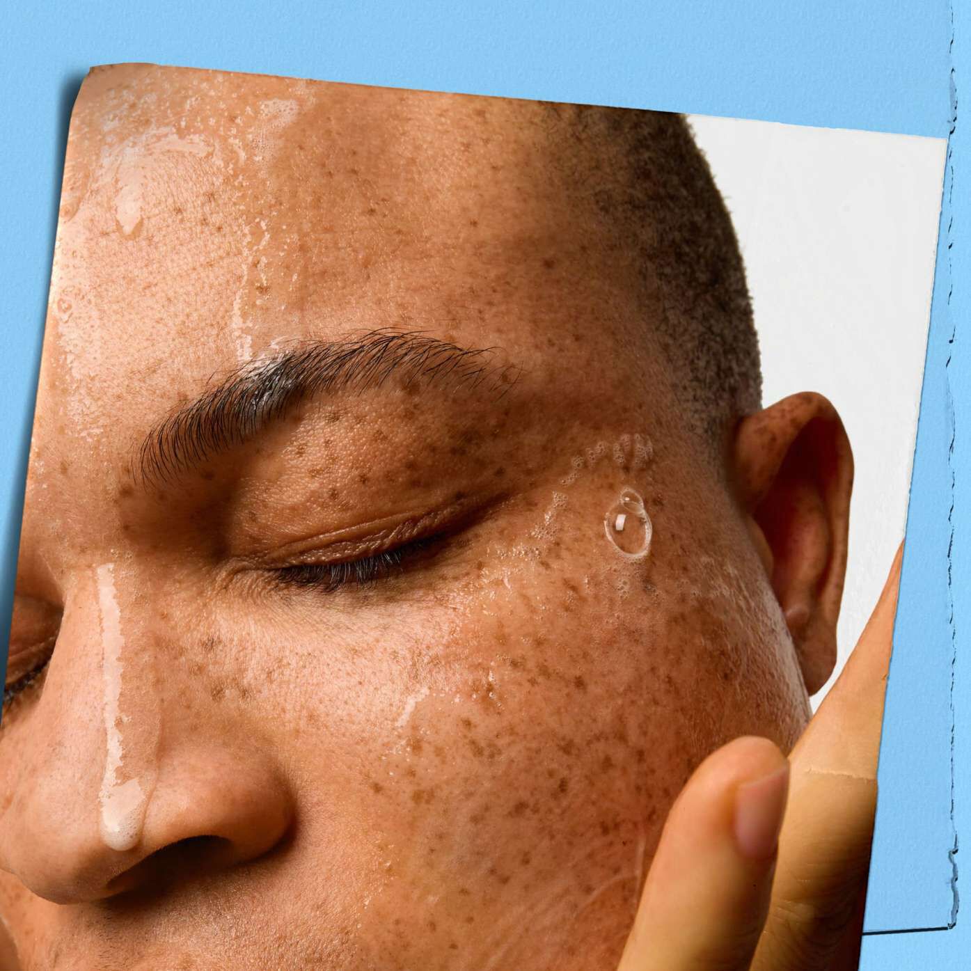 Close-up of a person with freckles and closed eyes, water droplets on their face. Their expression is calm and relaxed, with a blue background.