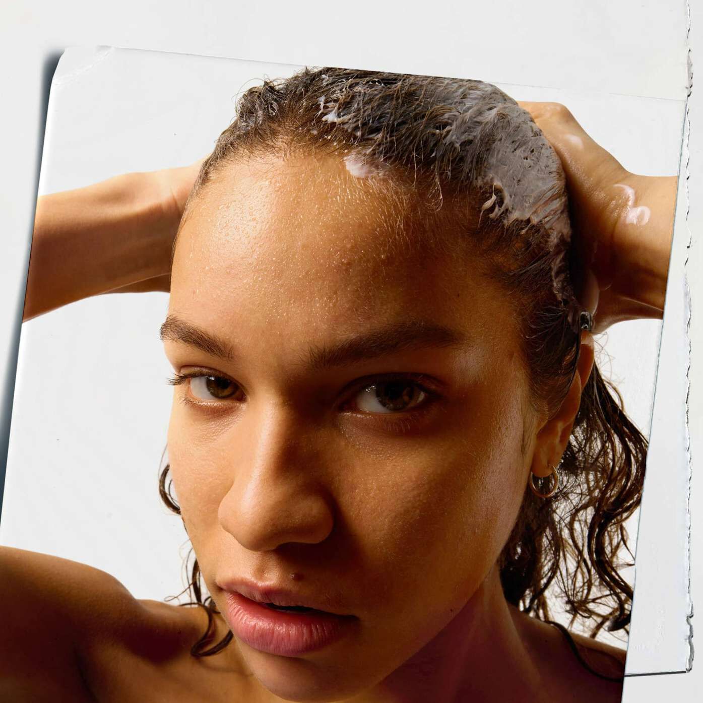 Close-up of a person with wet hair, hands massaging shampoo in. They look directly at the camera, conveying a focused and calm expression.