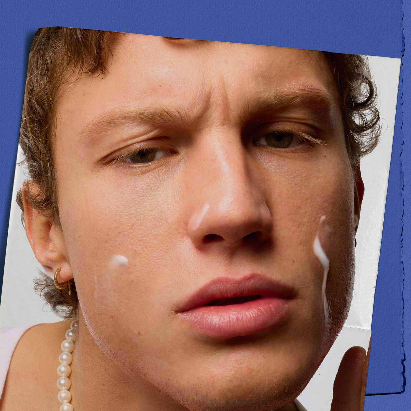A close-up of a person with a focused expression, examining their skin with lotion on their cheeks. They wear a pearl necklace and a gold earring, against a blue and white background.