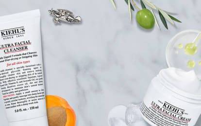 A flat lay of Kiehl's skincare products, including Ultra Facial Cleanser, Ultra Facial Toner, and Ultra Facial Cream, arranged on a marble surface with decorative elements like an orange, olives, and a small insect.