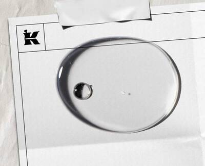 A close-up of a water droplet on textured paper, with a "K" logo in the top corner. The droplet reflects light and surroundings.