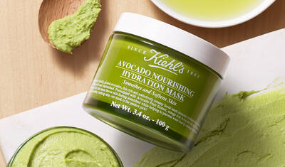 A green jar of Kiehl's Avocado Nourishing Hydration Mask, surrounded by avocado mask on a wooden surface and a small dish of olive oil.