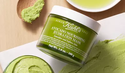 A green jar of Kiehl's Avocado Nourishing Hydration Mask, surrounded by avocado mask on a wooden surface and a small dish of olive oil.