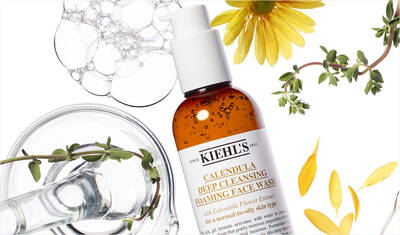 A bottle of Kiehl's Calendula Deep Cleansing Foaming Face Wash surrounded by flowers, herbs, and bubbles, on a light background.