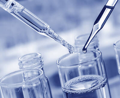 Close-up of two pipettes above a glass container, with droplets of liquid poised to be released into a transparent solution. The image has a cool blue tint.