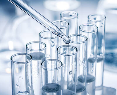 A pipette is dropping a liquid into a set of clear glass test tubes filled with colorless liquid, with an out-of-focus background of laboratory equipment.