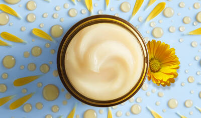 A top-down view of a creamy lotion in a circular jar, surrounded by yellow flower petals and water droplets on a light blue background.