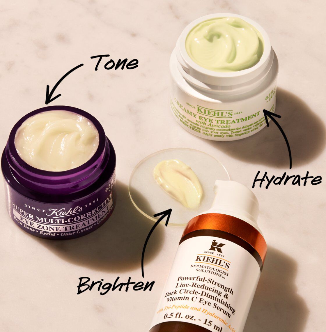 An overhead view of three skincare products on a marble surface: a purple jar labeled "Super Multi-Corrective Cream," a green jar labeled "Creamy Eye Treatment," and an orange bottle labeled "Powerful-Strength Dark Circle-Diminishing Vitamin C Eye Serum." Each product has a small amount of cream shown beside it, with arrows pointing to