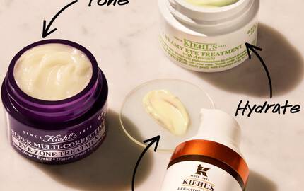 An overhead view of three skincare products on a marble surface: a purple jar labeled "Super Multi-Corrective Cream," a green jar labeled "Creamy Eye Treatment," and an orange bottle labeled "Powerful-Strength Dark Circle-Diminishing Vitamin C Eye Serum." Each product has a small amount of cream shown beside it, with arrows pointing to