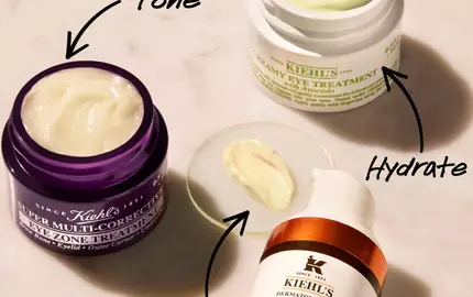 An overhead view of three skincare products on a marble surface: a purple jar labeled "Super Multi-Corrective Cream," a green jar labeled "Creamy Eye Treatment," and an orange bottle labeled "Powerful-Strength Dark Circle-Diminishing Vitamin C Eye Serum." Each product has a small amount of cream shown beside it, with arrows pointing to
