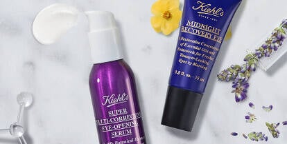 A flat lay of Kiehl's skincare products, including a green avocado eye cream, a lavender serum, and a purple moisturizer, arranged on a marble background alongside a dropper and an avocado slice.