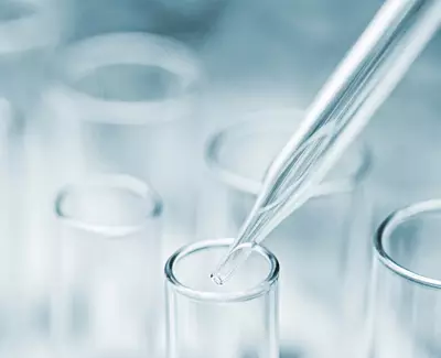 A close-up of a glass pipette about to dispense a drop into a test tube, with a blurred background of several test tubes.