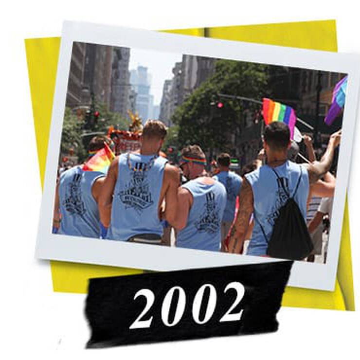 A group of men in blue tank tops participate in a Pride parade, holding rainbow flags, with city buildings in the background.