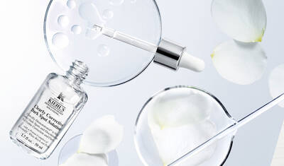 An elegant arrangement featuring a clear skincare serum bottle, a dropper, and glass petri dishes containing water droplets and flower petals, set against a soft, light background.