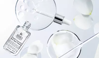 An elegant arrangement featuring a clear skincare serum bottle, a dropper, and glass petri dishes containing water droplets and flower petals, set against a soft, light background.