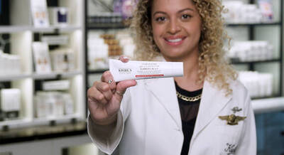 A person in a white coat is smiling and holding a tube of Kiehl's skincare product in a store filled with skincare items in the background.