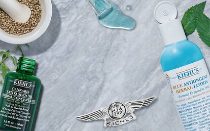 A flat lay arrangement of Kiehl's skincare products, including a green bottle of cannabis sativa seed oil and a blue bottle of hydrating lotion, surrounded by natural ingredients like herbs and oils on a marble surface.