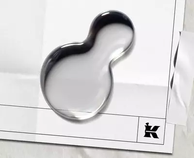 A shiny, abstract blob rests on white paper, featuring creases and a black 'K' logo in the bottom corner, creating a modern, artistic look.​