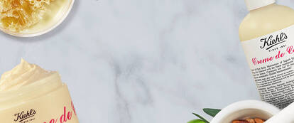 A flat lay of Kiehl's skincare products, including a jar of Creme de Corps and a bottle of Creme de Corps, surrounded by ingredients like honey, nuts, and olive branches on a marble surface.