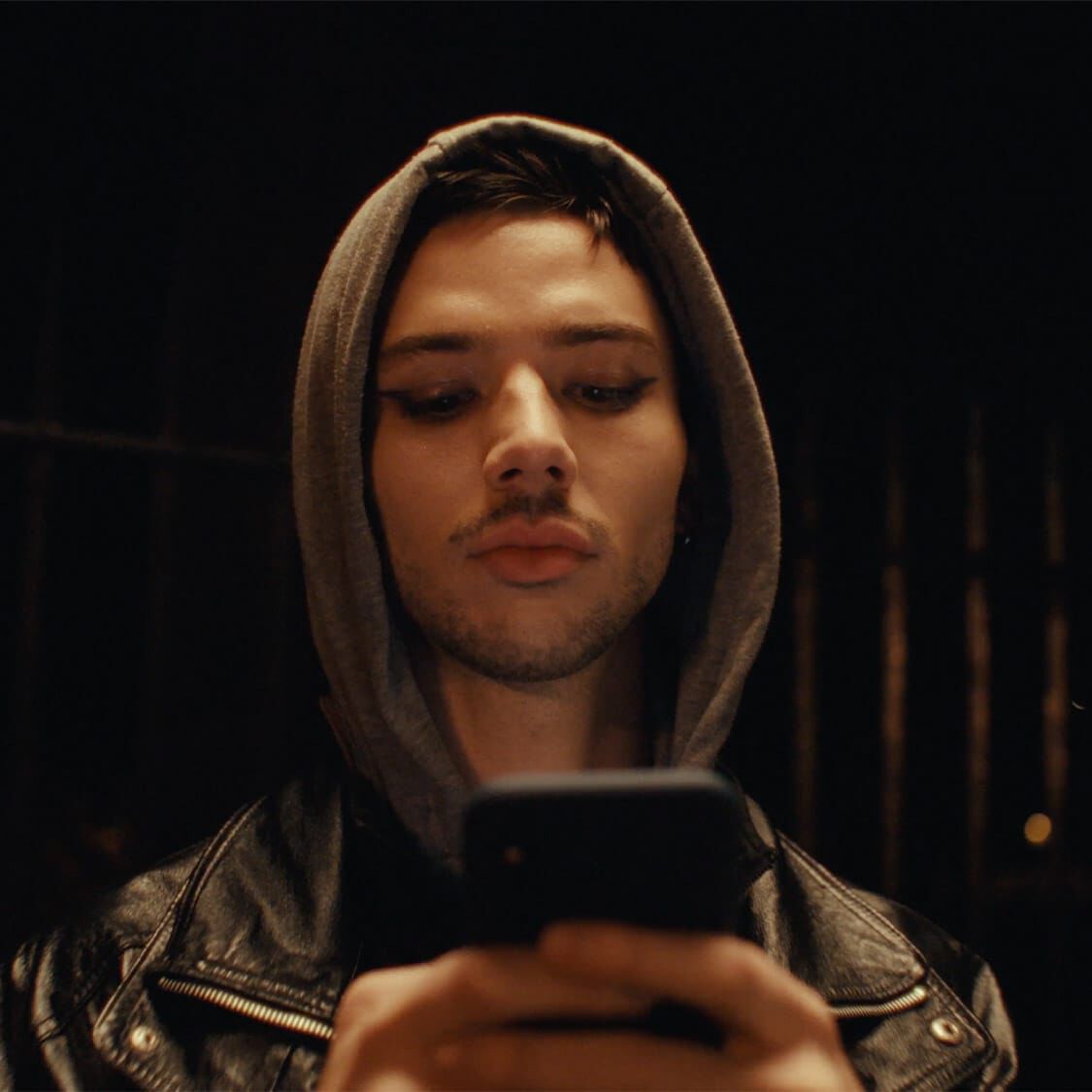 A figure in a hoodie and leather jacket stands in dim lighting, focused on their smartphone against a dark, blurred background.