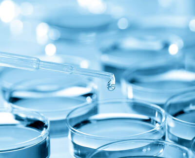 A dropper releasing a droplet into a row of transparent petri dishes filled with a blue liquid.