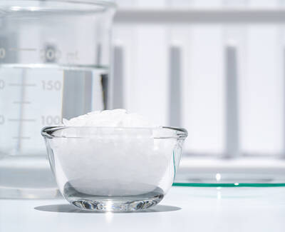 A clear glass bowl filled with white crystals sits on a table next to a measuring beaker with water. The background is bright and simple, featuring a white surface.