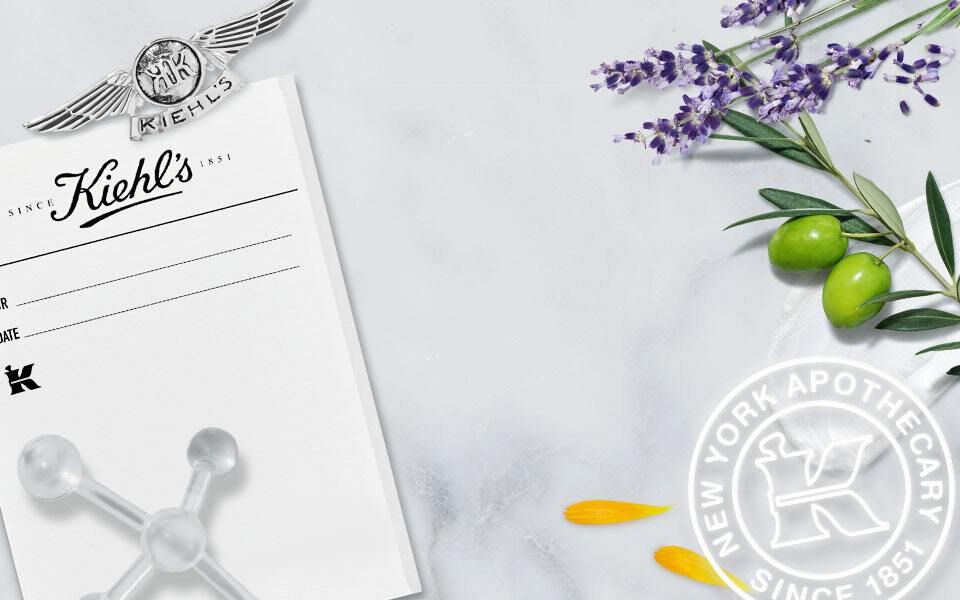 A collage featuring a vintage black-and-white photo of Kiehl's pharmacy staff in front of their store, accompanied by lavender, olive branches, and a white circular logo with "K" and "New York Apothecary." The background has a marbled texture.