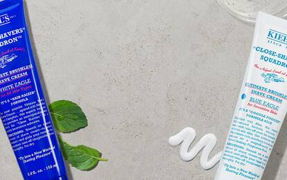 A flat lay of various Kiehl's men’s grooming products, including shaving creams and aftershaves, arranged on a light, textured surface. Mint leaves and a vintage style razor are also present, adding a fresh touch.