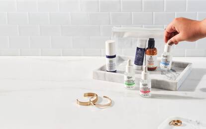 A hand holds a bottle of serum above a marble tray containing various skincare products and a small pouch, set against a light tiled background.
