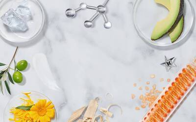 A flat lay of various natural ingredients and items on a marble surface, including flowers, herbs, an avocado, ice, and molecular structures, arranged symmetrically around the center.