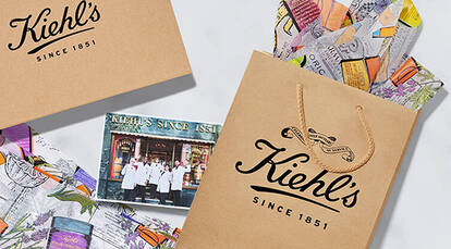 Three Kiehl's gift boxes with a beige design and white ribbons are displayed against a marbled background, alongside colorful artistic wrapping paper.