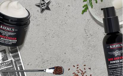 Alt text: A flat lay of Kiehl's skincare products, including jars of moisturizer and a bottle of serum, surrounded by coffee beans, a plant, and a small silver star on a light textured background.