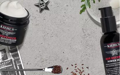 Alt text: A flat lay of Kiehl's skincare products, including jars of moisturizer and a bottle of serum, surrounded by coffee beans, a plant, and a small silver star on a light textured background.