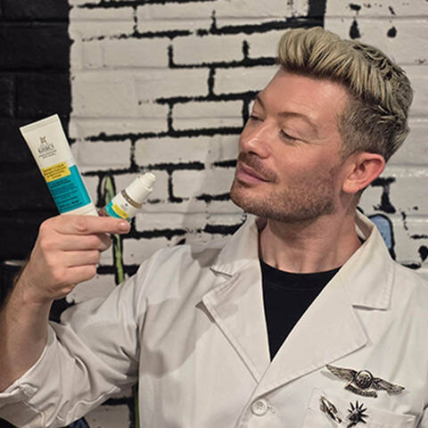 Kiehl's skin pro ain lab coat holds blemish skincare products, one in each hand, with a graffiti-style background of white and black bricks.