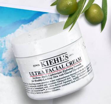 A jar of Kiehl's Ultra Facial Cream is displayed against a backdrop of icy mountains and snow, accompanied by olive branches and a small bowl of cream, emphasizing hydration and freshness.