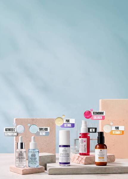 Kiehl's skincare product lineup on stone podiums, featuring five serums with ingredient labels for Activated C & Proxylane, Triple Acid Blend, Pure Retinol, Recombinant Collagen, and Pure Vitamin C.