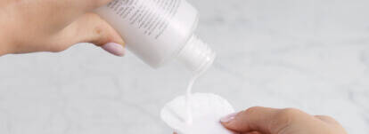 A person is pouring a liquid from a bottle onto a cotton pad, with a marble surface in the background.