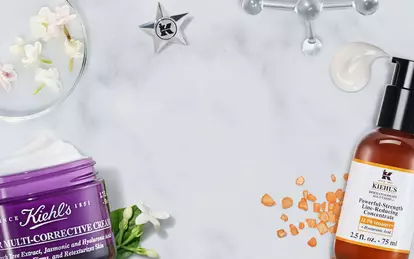 A flat lay of skincare products on a marble surface, featuring two bottles and a purple jar, surrounded by natural elements like flowers and orange peels.