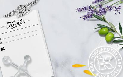 A collage featuring a vintage black-and-white photo of Kiehl's pharmacy with three staff members in white coats, alongside a marble background decorated with lavender, olive leaves, and petals, along with Kiehl's logo and text highlighting its history since 1851.
