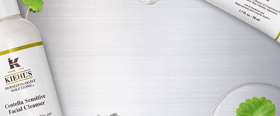 A flat lay featuring Kiehl's Cleansing Sensitive Facial Cleanser bottle alongside a cream tube, over a stainless steel background with botanical elements and laboratory glassware.