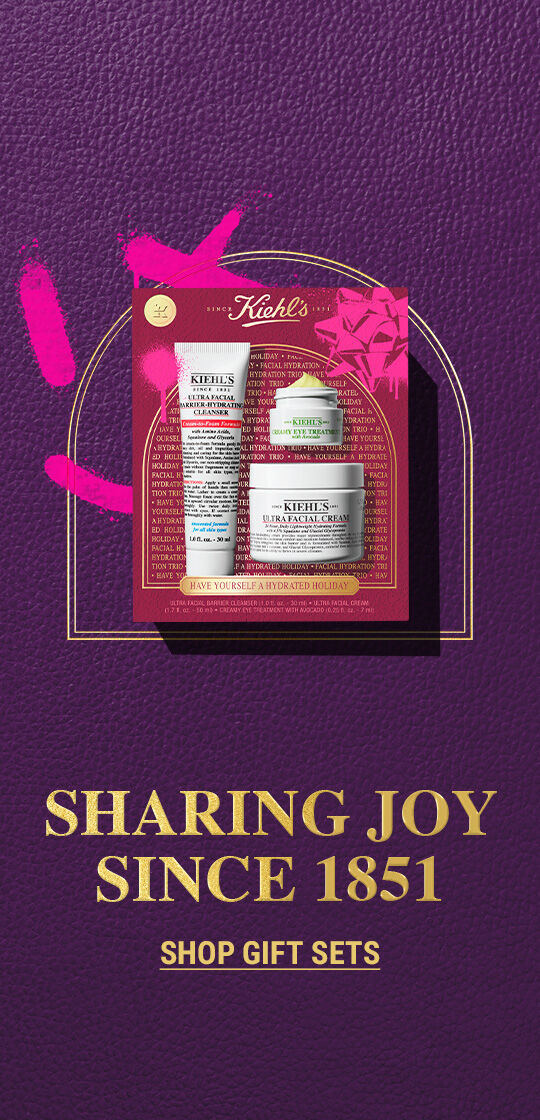 Kiehl's holiday gift set featuring a cleanser and facial cream, against a rich purple background with gold text. Slogan reads: Sharing Joy since 1951.