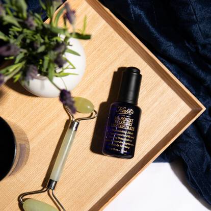 A flat lay image featuring a dark blue glass bottle of skincare serum on a wooden tray, accompanied by a jade roller and a small plant in a white pot, set against a black textured background.