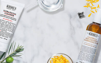 A flat lay of Kiehl's skincare products including Ultra Facial Cleanser, Calendula Herbal Extract Toner, and Facial Fuel, surrounded by natural ingredients like apricot, olive, marigold petals, coffee beans, and water on a marble surface.