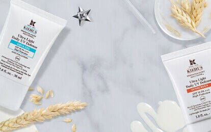 Alt text: A flat lay of Kiehl's skincare products, including two tubes of Ultra Light Daily UV Defense, alongside oat grains, a star charm, and a small vial, all on a marble surface.