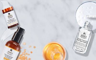 A flat lay of various skincare products on a marble surface, including serums in glass bottles and an orange liquid in a round dish, surrounded by small orange tablets.