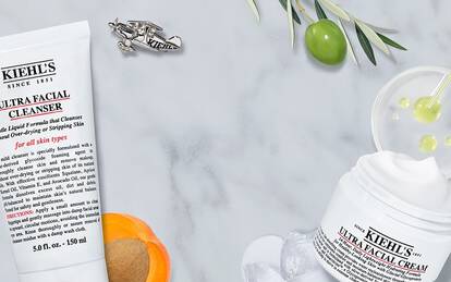 A flat lay of Kiehl's skincare products, including Ultra Facial Cleanser, Ultra Facial Toner, and Ultra Facial Cream, arranged with fresh orange slices and olive branches on a marble surface.