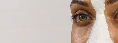 Close-up of a woman's face with a facial mask applied on one side, highlighting her eye and skin texture.