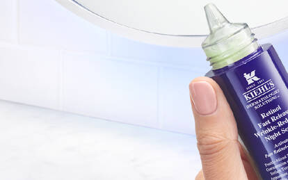 A hand holding a bottle of Kiehl's skincare product, with other Kiehl's items and a towel visible in the background. The setting is bright and clean, likely in a bathroom.